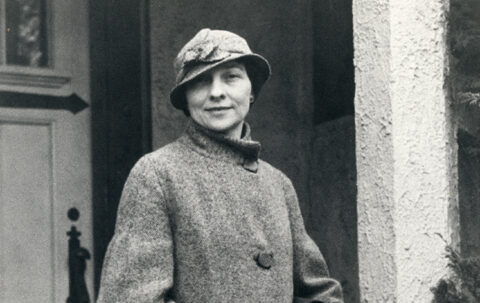 Black and white photograph of Elizebeth Friedman standing in front of possibly her residence during her career with U.S. Coast Guard. Elizebeth is carrying a bag and wearing a long coat and hat.