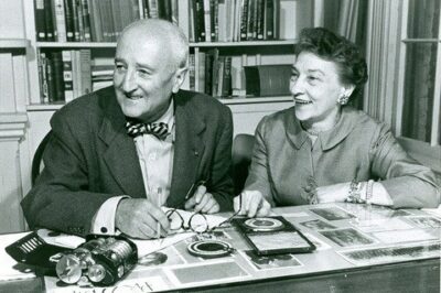 Black and white image of Elizebeth Friedman and husband William sitting at a desk with a full bookshelf behind them. Image taken later in life after years of valuable intelligence service for the nation. Friedman was a pioneering code-breaker for the Coast Guard during the prohibition era and World War II.