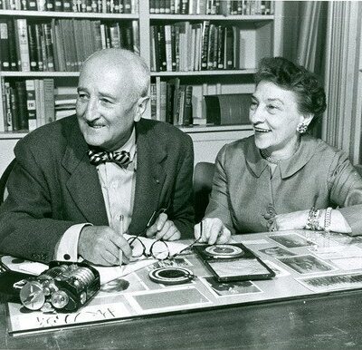 Black and white image of Elizebeth Friedman and husband William sitting at a desk with a full bookshelf behind them. Image taken later in life after years of valuable intelligence service for the nation. Friedman was a pioneering code-breaker for the Coast Guard during the prohibition era and World War II.