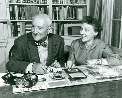 Black and white image of Elizebeth Friedman and husband William sitting at a desk with a full bookshelf behind them. Image taken later in life after years of valuable intelligence service for the nation. Friedman was a pioneering code-breaker for the Coast Guard during the prohibition era and World War II.