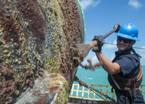 Photo: A Coast Guard crew member wields a large heavy scraping tool while cleaning off the bottom of a buoy.