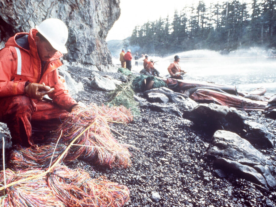A color photograph of a Coast Guard member as they check the oil recovery mesh as others steam blast rocks and shorelines soak by with crude oil from the spilled tanker Exxon Valdez.