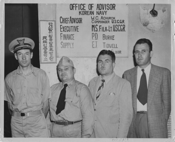 Photo: Four men standing in front of a sign “Office of Advisor, Korean Navy. Chief Advisor: W.C. Achurch, Commander U.S.C.G.R, Executive: M.S. Fila-Lt. U.S.C.G.R.; Finance: P.D. Burke; Supply: E.J. Tidwell.”