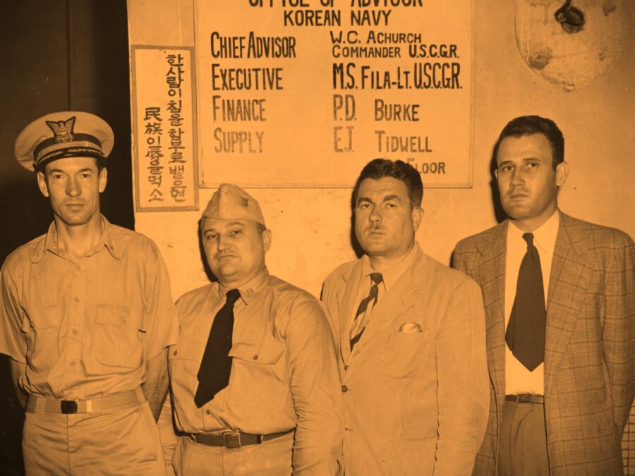 Photo: Four men standing in front of a sign “Office of Advisor, Korean Navy. Chief Advisor: W.C. Achurch, Commander U.S.C.G.R, Executive: M.S. Fila-Lt. U.S.C.G.R.; Finance: P.D. Burke; Supply: E.J. Tidwell.”