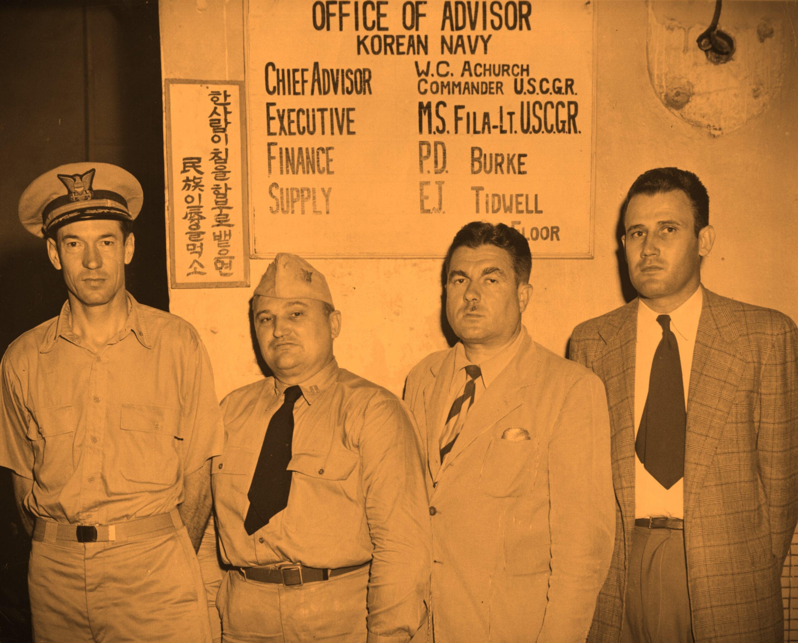 Photo: Four men standing in front of a sign “Office of Advisor, Korean Navy. Chief Advisor: W.C. Achurch, Commander U.S.C.G.R, Executive: M.S. Fila-Lt. U.S.C.G.R.; Finance: P.D. Burke; Supply: E.J. Tidwell.”