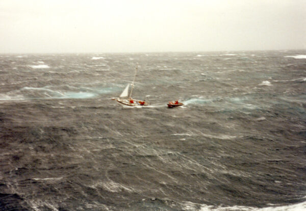 Photo: Distant aerial view of inflatable rescue boat approaching a sailboat in rough seas.