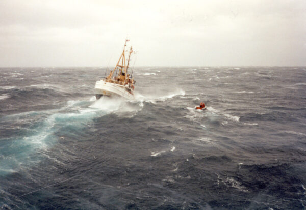 Photo: Color photograph of cutter Tamaroa plunging through heavy seas during the Perfect Storm, with a small orange life raft or boat visible to the right.