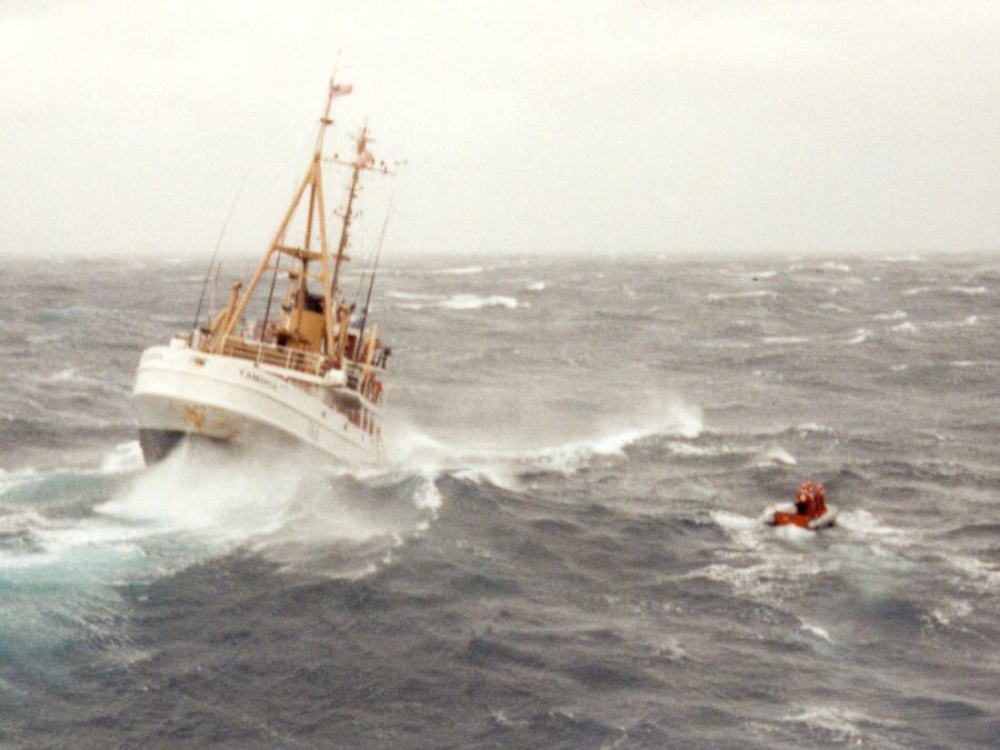 Photo: Color photograph of cutter Tamaroa plunging through heavy seas during the Perfect Storm, with a small orange life raft or boat visible to the right.