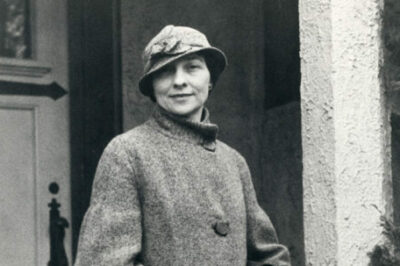 Black and white photograph of Elizebeth Friedman standing in front of possibly her residence during her career with U.S. Coast Guard. Elizebeth is carrying a bag and wearing a long coat and hat.