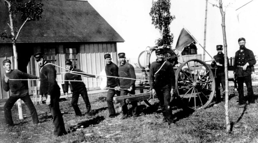 Black & white photograph of unidentified US Life Saving Service keeper & seven crew members in standard uniforms pulling a beach cart. Station building in foreground.