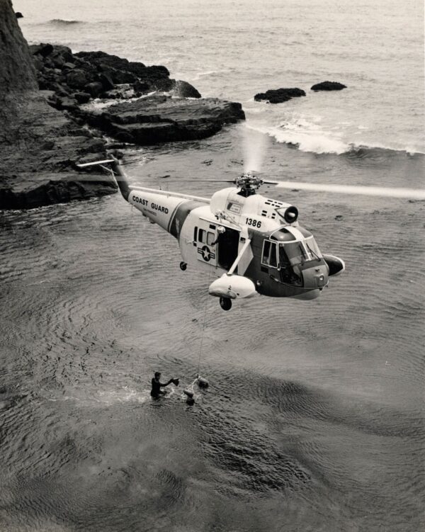 Photo: Black-and-white photograph of an HH-52 helicopter hovering over a SARWET member in the water near rocky coastline during training.