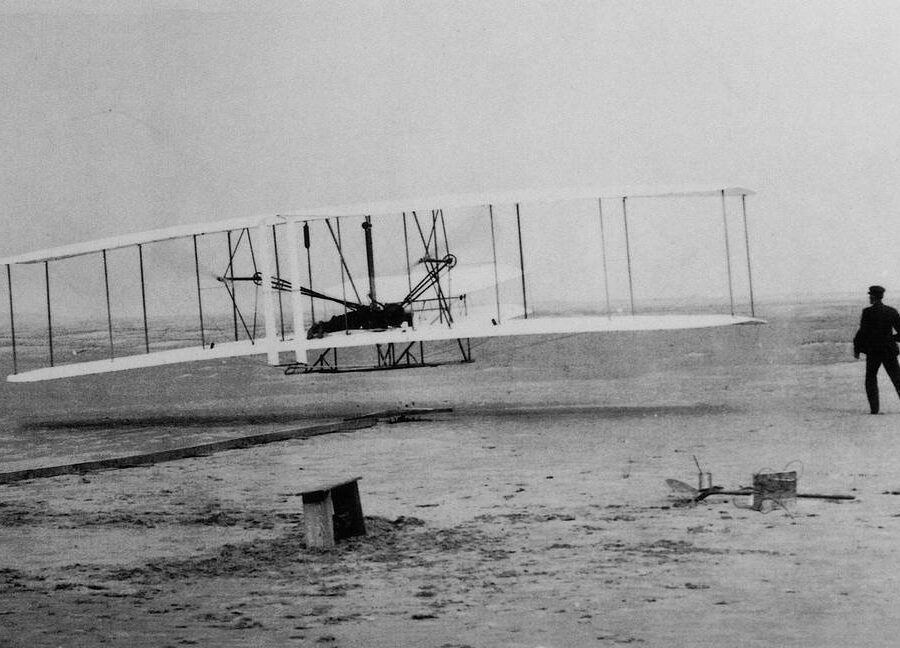 Black and white photography of Orville Wright at the controls and Wilbur Wright running alongside, the Wright Flyer makes its first flight at Kitty Hawk, NC, Dec 17, 1903.