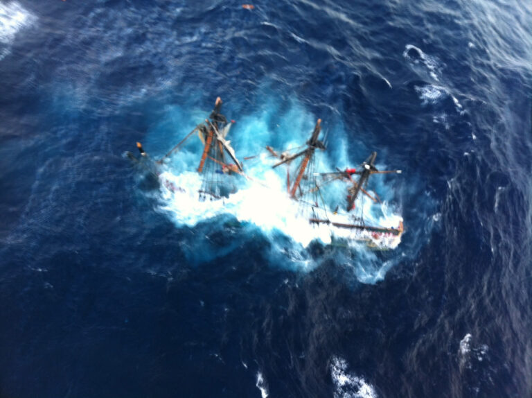 Photo: Aerial view of HMS Bounty sinking, decks already submerged while masts extend from the water.