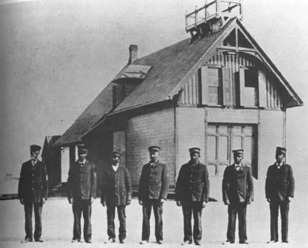 Photo of seven men in uniform posing in a row in front of the Pea Island Life-Saving Service Station.