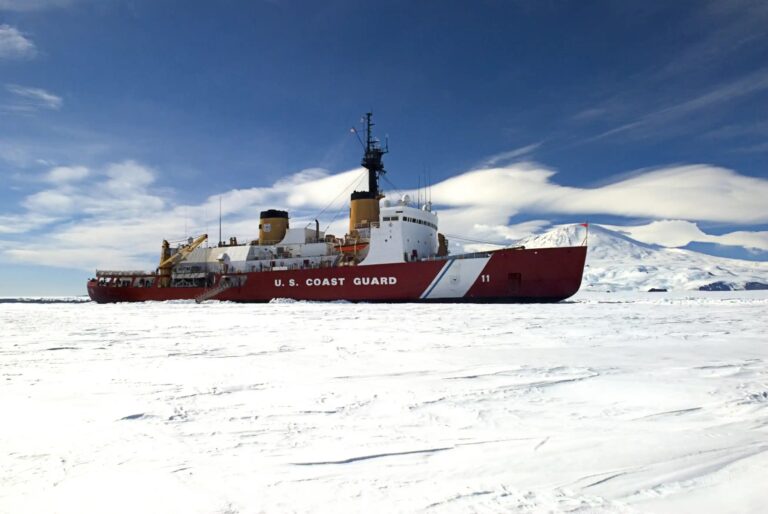 Photo: Icebreaker Polar Sea under a blue sky, with ice in foreground and snowy mountain in the background.