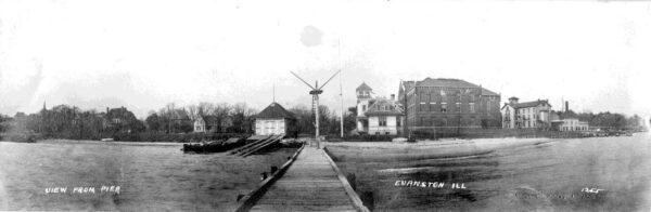 Photograph of the Station Evanston complex, with the original 1871 boathouse on the left, station wreck pole with dock at center, expanded 1877 station building at right of center, Fisk Hall next to the station, and the remaining structures are NWU buildings.