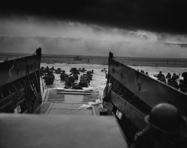 Photo taken from perspective of troop transport on the shores of Normandy where soldiers exiting the transport head through the water to the beach.