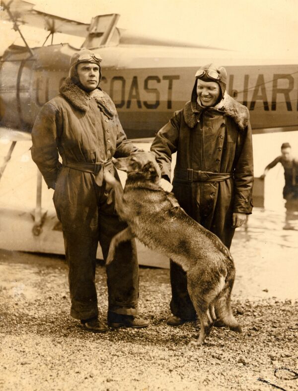 Two pilots pose in front of a Coast Guard sea plane along with their dog.
