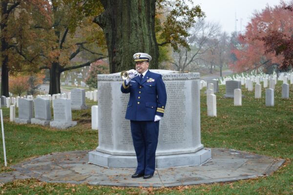 Photo: Uniformed Coast Guard officer blowing a bugle with gravesite in background.