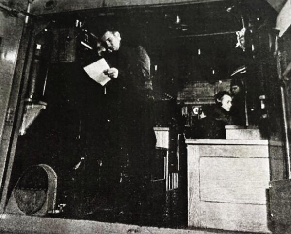 Photo: Inside a Coast Guard HF-DF Net Truck, a man reads from a paper while another seated man listens to headphones.