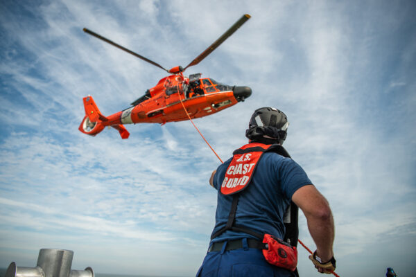 Color photograph of Petty Officer 3rd Class Hayden Falkenberry, a machinery technician at Coast Guard Station New Orleans, conducts helicopter hoist training. Photo taken low to the ground with view of Falkenberry holding a hoist that leads to a red Coast Guard helicopter.