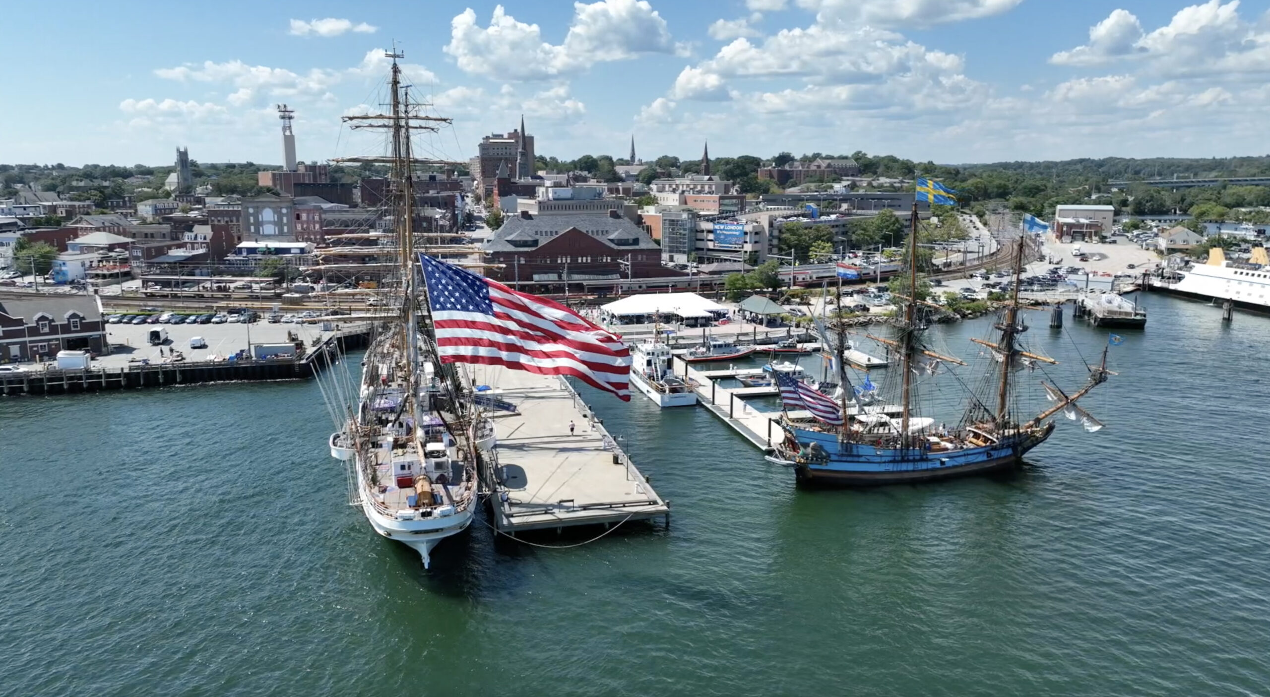 Color (water view - Aerial) photograph of Keel Laying Ceremony and Construction Site with America's Tall Ship the Coast Guard Cutter Eagle moored pierside. A very large Americal flag is flying.