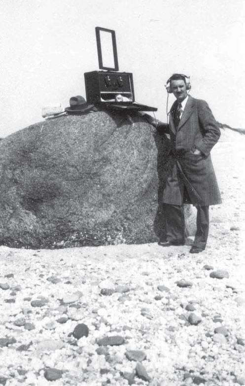 Photo: Man with headphones standing next to a radio with antenna sitting on a large boulder.