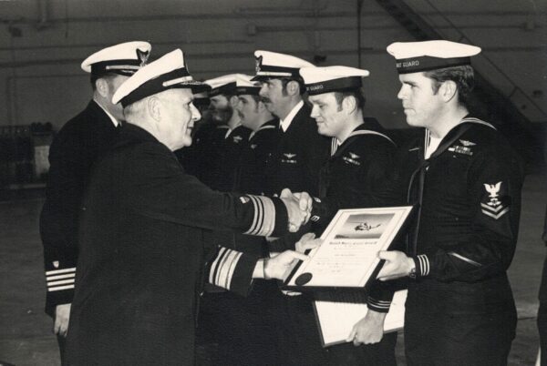 Photo: Black-and-white photograph of Rear Admiral Mark Whalen and Captain Charles Larkin presenting SARWET designations to team members in a hangar.