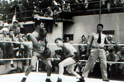 Photo in black and white: Two boxers in a ring with referee Heavyweight boxer and Coast Guard officer, Jack Dempsey, in the background are sailors looking on.