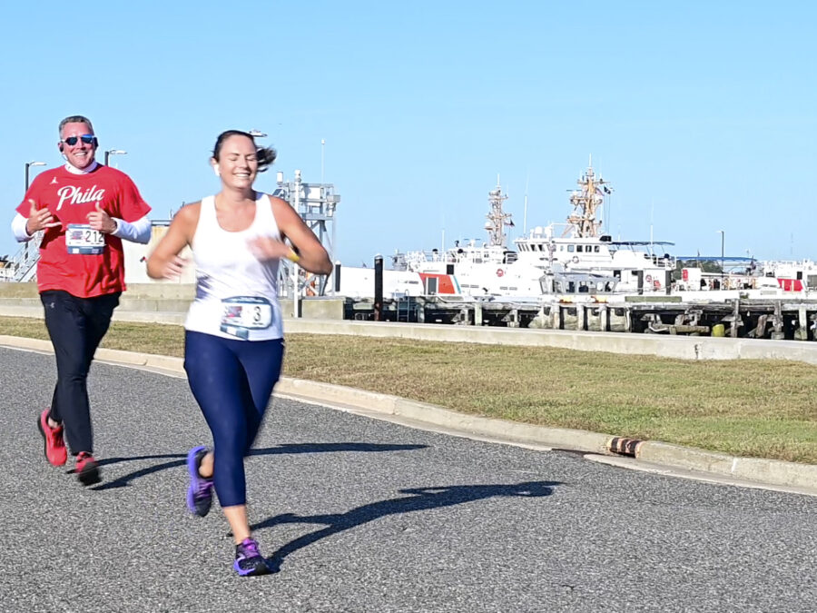 Color photograph of runners participating in the inaugural Always Ready 5K, Oct. 15, 2022, during the on-base segment at Training Center Cape May. Three Coast Guard cutters are moored behind the runners.