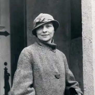 Black and white photograph of Elizebeth Friedman standing in front of possibly her residence during her career with U.S. Coast Guard. Elizebeth is carrying a bag and wearing a long coat and hat.